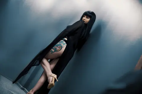 Elegant woman in a flowing black gown with leg tattoos posing against a moody blue wall.