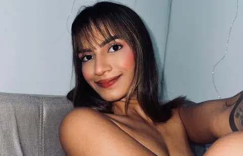 Smiling young woman with brown hair and makeup relaxing on a couch in a bright indoor setting.