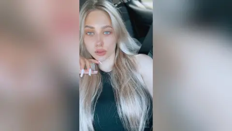Blonde woman with blue eyes wearing a black tank top sitting in a car with a natural, serene expression.