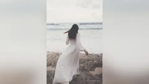 StacyHarland stands on a sandy beach wearing a long white dress with long sleeves, looking out toward the ocean waves.