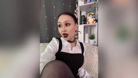 Young woman in a white shirt and black tank top looking surprised in a cozy bedroom setting.