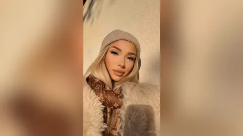 Stylish woman in a beige beanie and fuzzy white coat posing outdoors in warm sunlight.