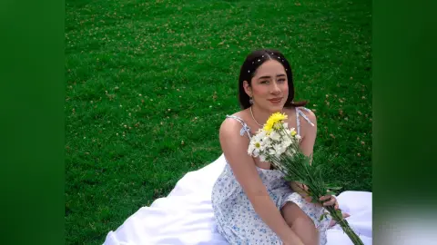 IsabellaYork — Smiling young woman in a white floral sundress holding a bouquet of daisies on a picnic blanket in a green field.