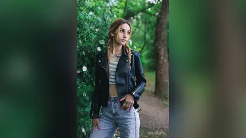 Stylish young woman with braided hair wearing a black leather jacket and jeans in a lush green forest setting.