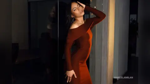 MarcelaMilan poses in a tight, long-sleeved red dress with her hand on her head against a dark glass window.