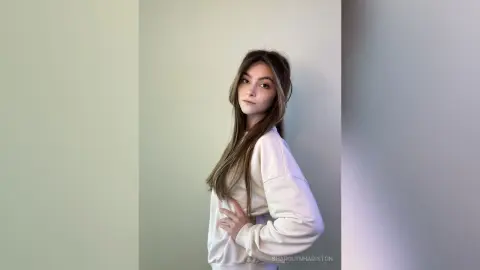 Young woman in a white sweatshirt posing against a minimalist light background with a calm expression.