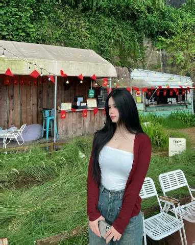 Young woman in a white tank top and jeans relaxing at a rustic outdoor cafe with lush green foliage.