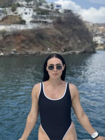 Athletic woman in a black one-piece swimsuit and sunglasses relaxing by the ocean on a sunny day.
