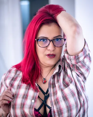 Red-haired woman in a plaid shirt and black lace top posing indoors with a confident mood.