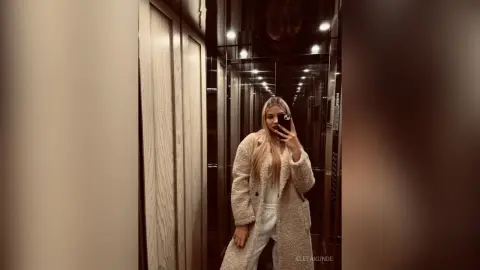 AletaKunde poses for a mirror selfie in an elevator, wearing a long, fuzzy cream-colored coat and white pants with her long blonde hair down.