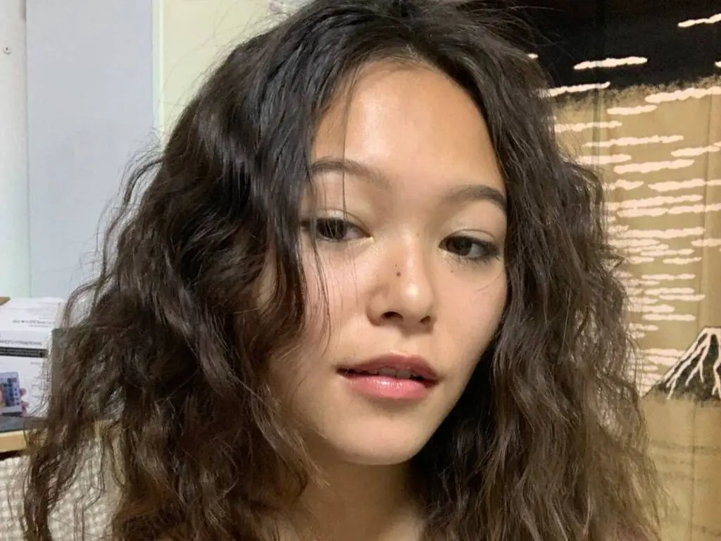 Close-up portrait of a young woman with wavy brown hair looking calmly at the camera in a bright indoor setting.