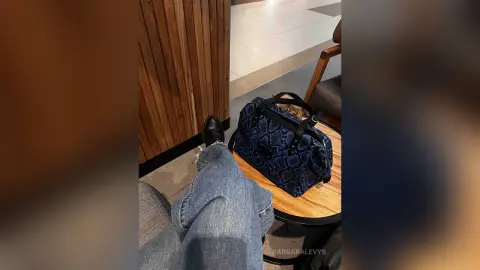 Person in blue jeans and black boots sitting next to a navy snakeskin print handbag in a modern interior.