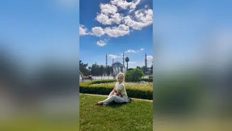 Blonde woman in a white top sitting on green grass before the Blue Mosque under a bright blue sky.