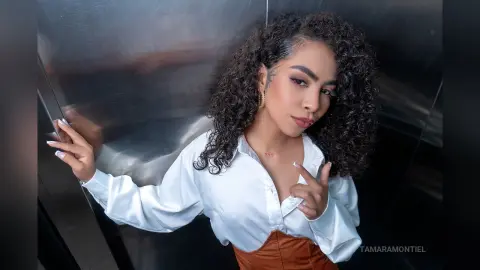Curly-haired woman in a white button-down shirt posing confidently against a metallic silver background.