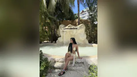 Aleja sits on a stone ledge by a small waterfall, wearing a light yellow bikini and black sandals.