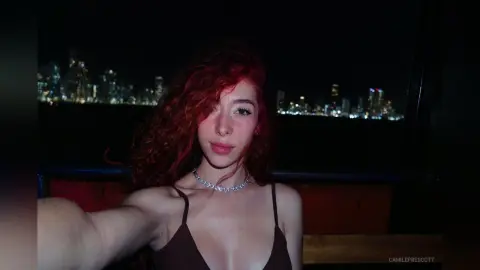 Young woman with curly red hair wearing a dark tank top and silver necklace against a night city skyline.