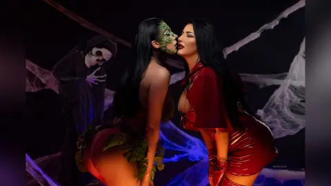 MissyJolie poses in a green leaf bikini and dark hair, leaning in to kiss a skeleton figure against a backdrop of spiderwebs.