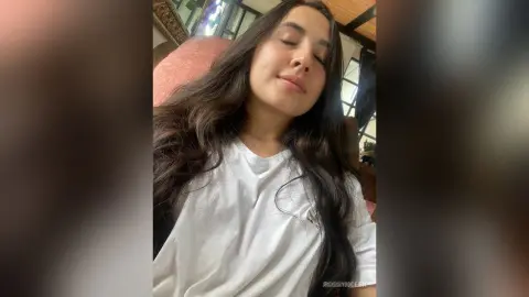 Smiling young woman with long dark hair wearing a white t-shirt in a bright indoor setting.