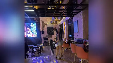 Stylish woman in a grey mini dress standing in a modern, dimly lit lounge with ambient blue lighting.