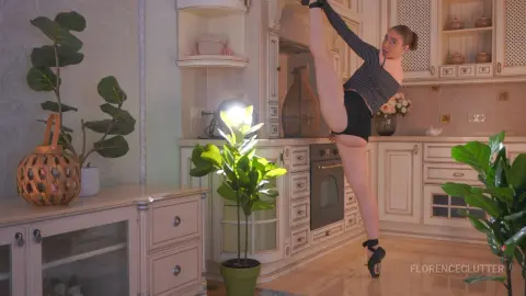 FlorenceClutter stands in a kitchen, lifting one leg high up toward her head while wearing a black striped top and black shorts.