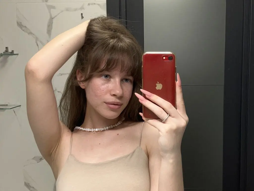Young woman with bangs in a beige camisole and pearl necklace taking a mirror selfie in a modern bathroom.