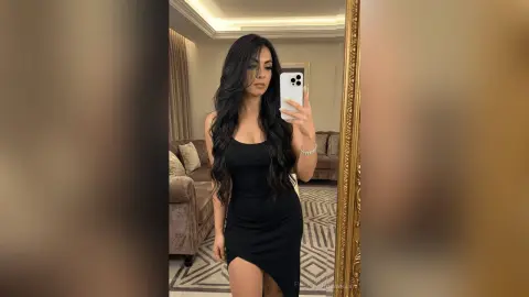 Woman with long wavy hair in a black mini dress taking a mirror selfie in a modern living room.