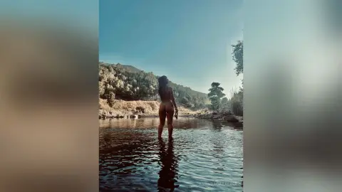 Curvy woman in a bikini standing in a tranquil river surrounded by lush green mountains under a clear sky.