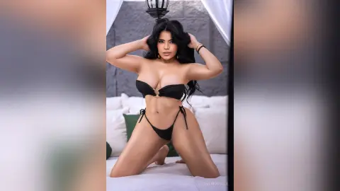 DayanaHille poses on a white bed wearing a black string bikini, running her hands through her long dark hair.