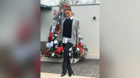AnaMathers smiles while wearing a grey blazer over a black sweater and white shirt with black leggings and black boots.