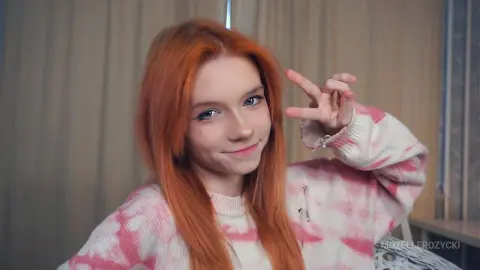 Smiling redhead woman in a pink tie-dye sweater making a peace sign in a cozy indoor setting.