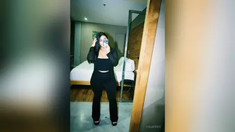 Young woman in a black crop top and trousers taking a mirror selfie in a modern bedroom.