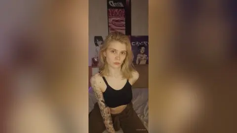 Blonde woman in a black crop top sitting in a bedroom with anime posters, featuring a relaxed mood.