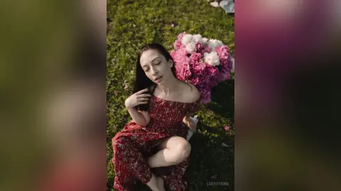 Dreamy portrait of a woman in a red floral dress sitting on green grass next to a large pink bouquet.