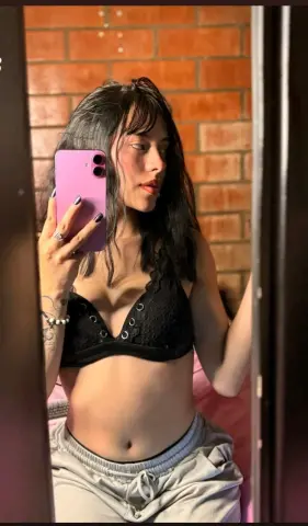Young woman in a black lace bralette taking a mirror selfie against a brick wall background.