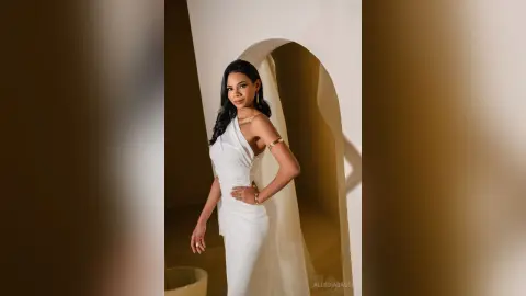 Elegant woman in a white satin gown posing in a minimalist arched doorway with soft, warm lighting.