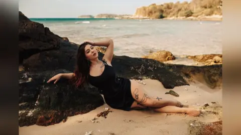 Tattooed woman in a black dress relaxing on a sandy beach by rocky cliffs under a bright sky.
