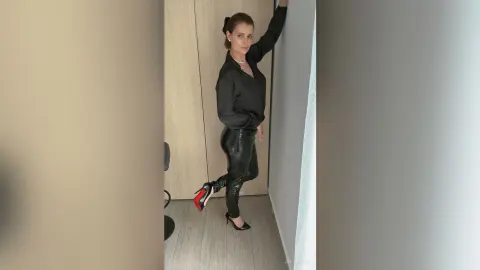 Elegant woman in black blouse and leather pants posing confidently in a modern minimalist room.