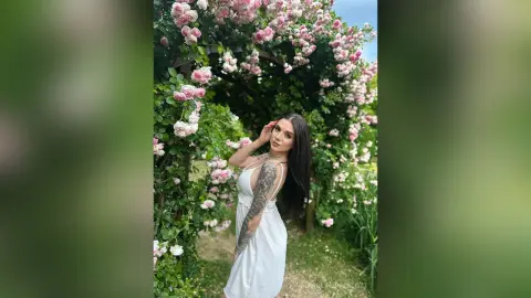 Young woman in a white summer dress smiling amidst a lush pink rose garden archway.