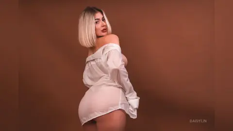 Blonde woman in a white sheer shirt posing against a brown studio background with a moody aesthetic.