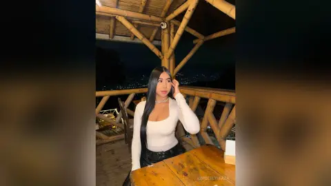 KimberlyIsaza poses in a white long-sleeve top and black pants, running her hands through her long dark hair in a bamboo hut at night.