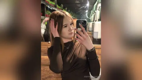 Young woman with long brown hair wearing a black long-sleeve top taking a mirror selfie in a dimly lit room.