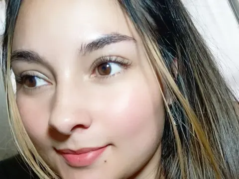 Close-up portrait of a smiling young woman with long brown hair and natural makeup in soft indoor lighting.