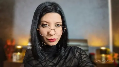 Woman with dark hair and gold glasses wearing a black sheer patterned top in a warm, moody indoor setting.