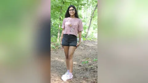 Smiling young woman in a floral graphic t-shirt and black denim shorts standing in a sunny forest setting.