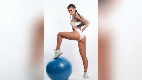 Tattooed woman in white fitness wear posing with a blue exercise ball in a bright studio setting.