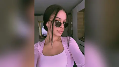 AriaVex wears dark sunglasses and a light pink tank top and cardigan, looking off to the side with her black hair styled down.