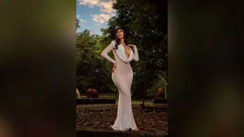 Elegant woman in a white long-sleeve evening gown posing in a lush green garden setting.
