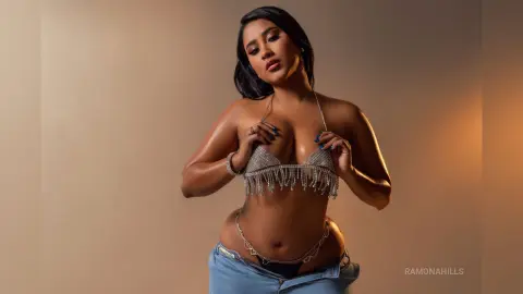Confident woman in a sparkling silver sequin bikini top and denim jeans posing against a neutral studio background.