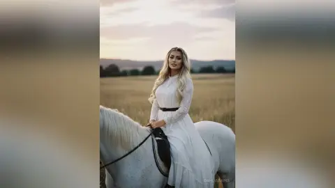 Elegant woman in a long white lace dress riding a white horse through a dreamy golden field at sunset.