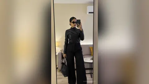 Stylish woman in a black turtleneck and wide-leg trousers taking a mirror selfie in a modern living room.